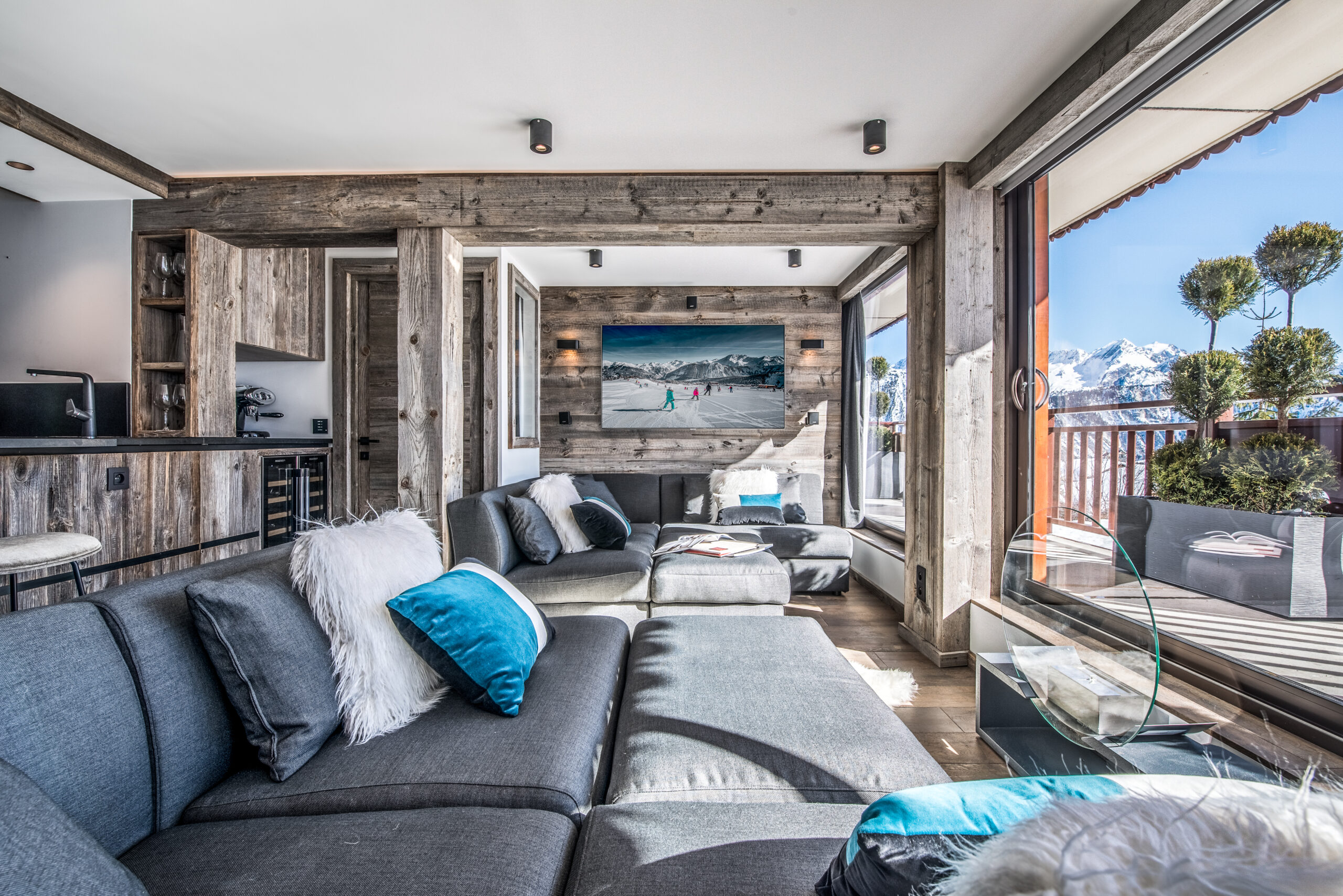 Modernes Wohnzimmer mit grauem Sofa und Altholz-Wänden aus Mareiner Zermatt Gris. Große Fenster bieten freien Blick auf verschneite Berge. Stilvolle Kombination aus rustikalem Holz und elegantem Design.