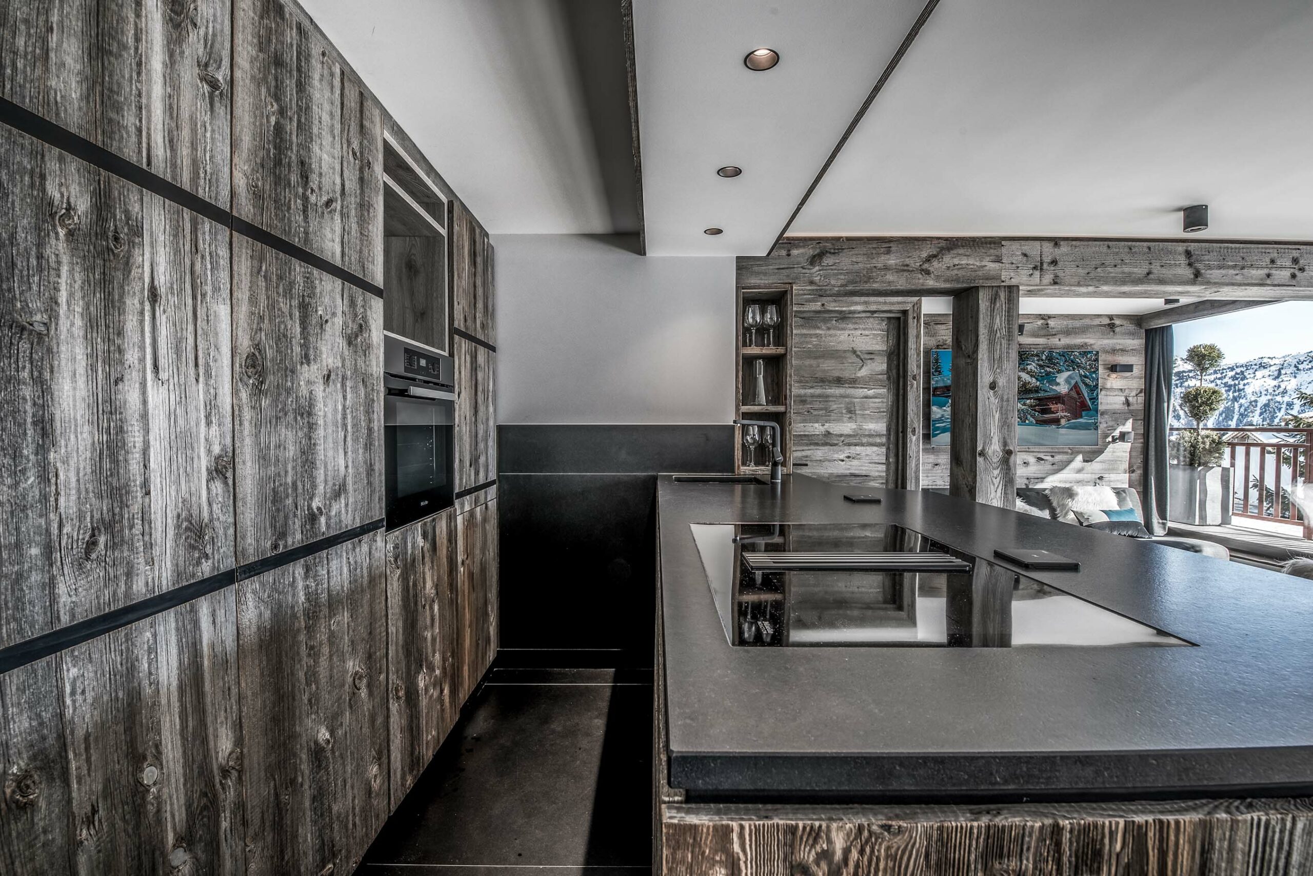 Moderne Hotelküche mit Fronten und Wandverkleidung aus Mareiner Altholz Zermatt Gris, kombiniert mit schwarzer Stein-Arbeitsplatte, hochwertiger Ausstattung und Blick auf die verschneite Berglandschaft.