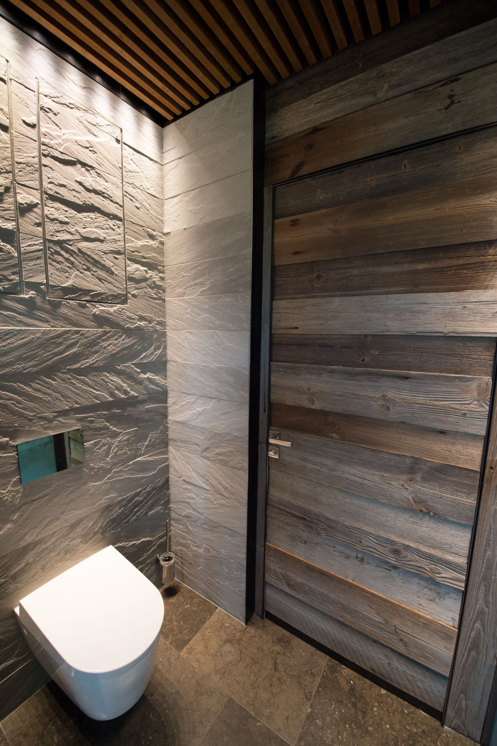 Modernes WC mit strukturierter Steinwand und Tür aus Holz, kombiniert mit dunklem Natursteinboden und Holzlattendecke.