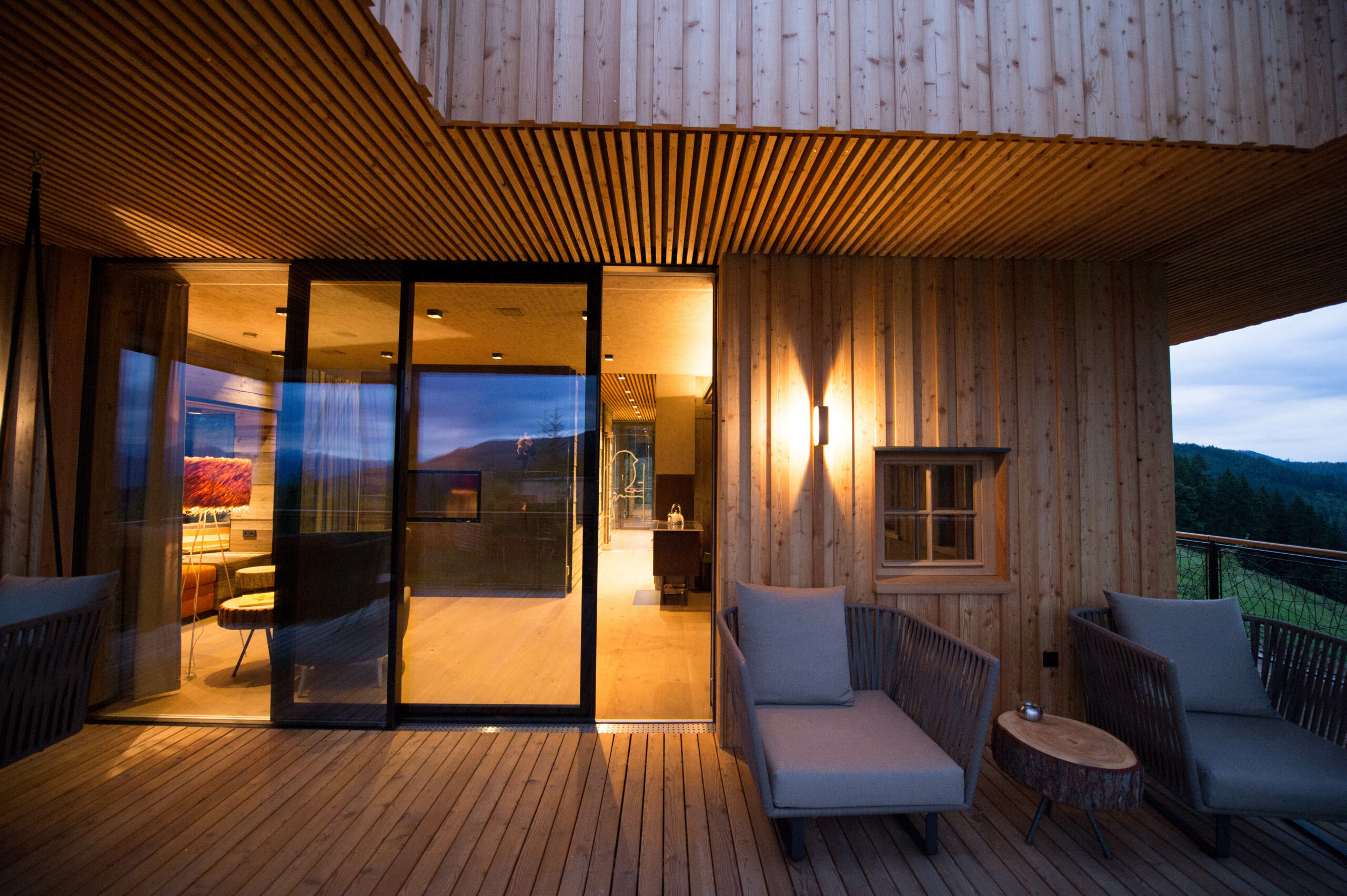 Gemütliche Terrasse eines Chalets am Abend, mit vertikaler Holzfassade und Decke aus Mareiner Holz Piz Bernina, stilvollen Outdoor-Möbeln und Blick in die Berglandschaft.