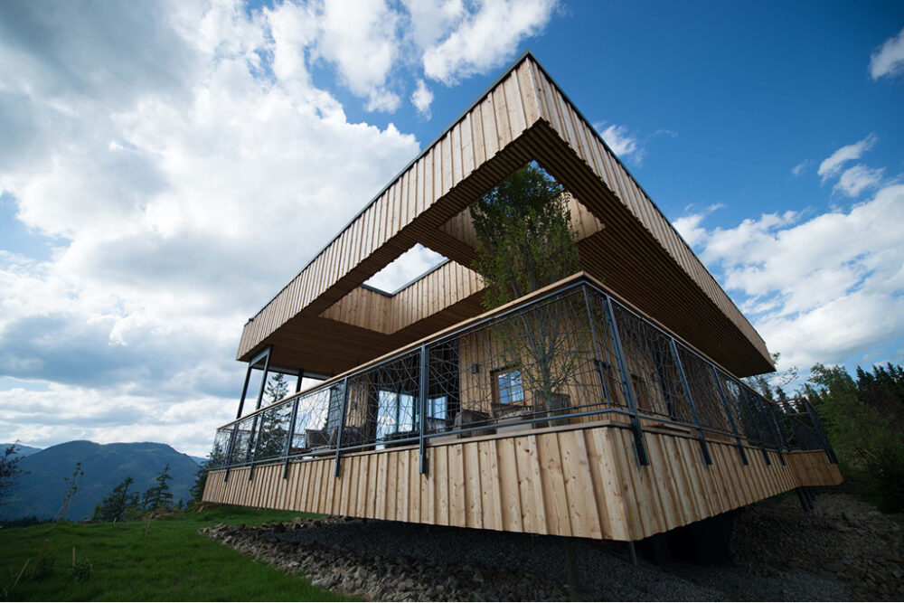 Modernes Holzchalet mit auffälliger Architektur auf Stelzen, verkleidet mit vertikalen Holzlamellen aus Mareiner Holz Piz Bernina, inmitten alpiner Landschaft unter blauem Himmel.