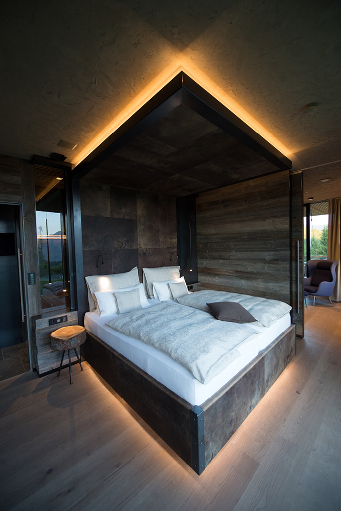 Stilvolles Schlafzimmer mit indirekter Beleuchtung, Holzwänden und rustikalem Bett mit Stahlrahmen. Naturmaterialien und moderne Elemente schaffen eine gemütliche Atmosphäre mit Blick ins Freie.