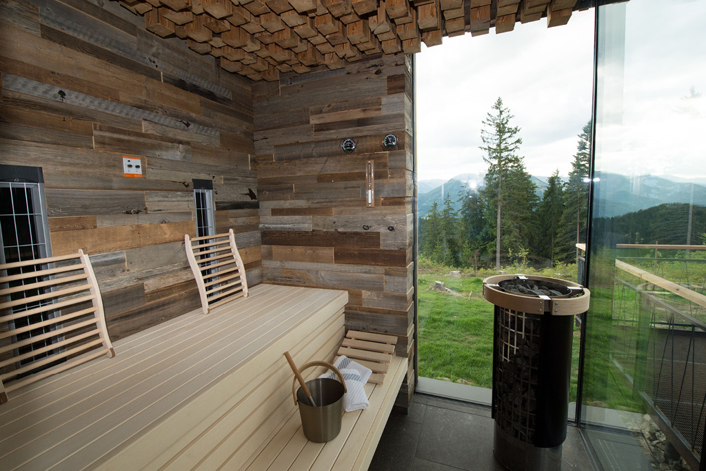 Moderne Sauna mit Altholzverkleidung (Mareiner Zermatt Gris) und großer Glasfront, die den Blick auf Wald und Berge freigibt. Holzbänke, Saunaofen und Aufgusskübel im Vordergrund.