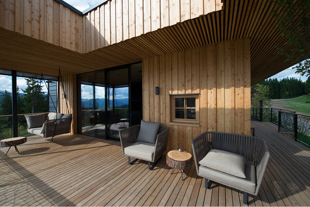 Überdachte Holzterrasse eines Chalets mit gemütlichen Sitzmöbeln, Wänden und Decke aus Mareiner Piz Bernina Holz, Blick ins Grüne und große Glasfronten zur Verbindung von Innen- und Außenbereich.