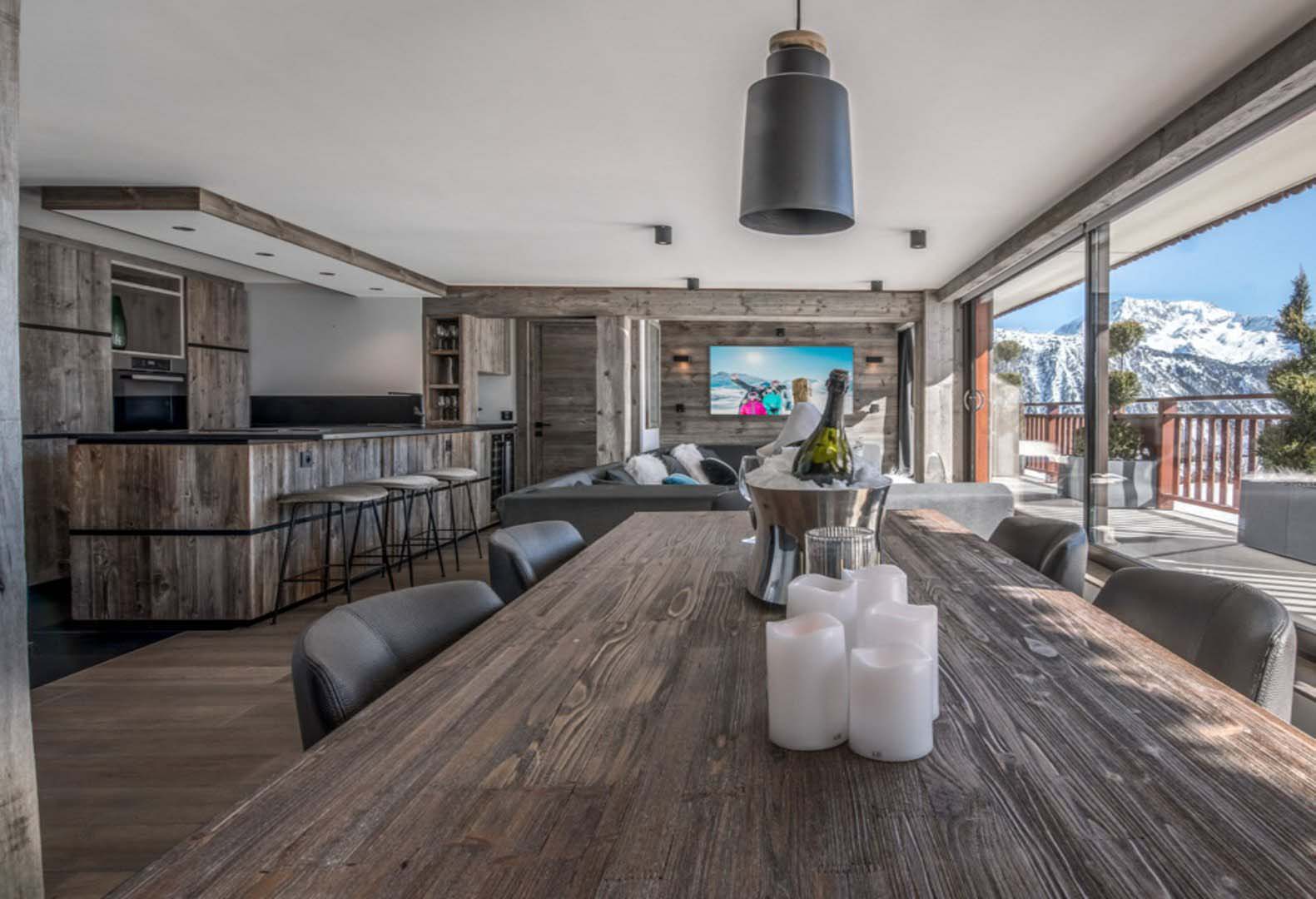 Offener Wohn- und Essbereich mit Esstisch aus Altholz, Küche, Sofa und Bergblick. Wände und Möbel aus Mareiner Altholz Zermatt Gris schaffen ein edles, gemütliches Ambiente mit Sicht auf verschneite Alpen.