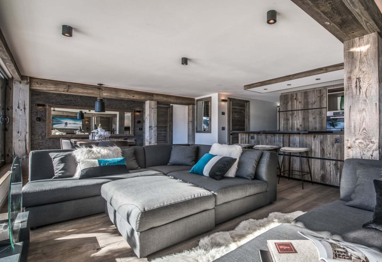 Heller Wohn- und Essbereich mit grauem Sofa, Esstisch und Küche, gestaltet mit Mareiner Altholz Zermatt Gris. Große Fenster eröffnen den Blick auf die Berge, modernes Ambiente trifft rustikale Holzstruktur.