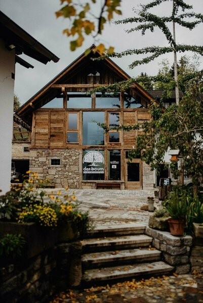 Vorderansicht des Seminarzentrums der Baum mit Naturstein- und Altholzfassade, großer Glasfront, Treppe im Vordergrund, herbstlicher Bepflanzung und einem Firmenschild am Gebäude.