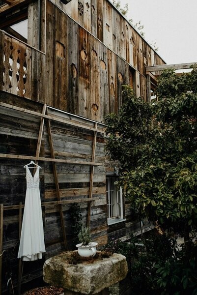 Hölzerne Fassade des Seminarzentrums der Baum aus Mareiner Altholz, davor ein weißes Hochzeitskleid an der Wand und ein alter steinerner Brunnen im Garten mit Pflanzen.