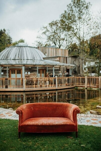 Ansicht des Seminarzentrums der Baum mit Holzfassade aus Mareiner Altholz, Teich und rotem Sofa im Vordergrund, eingebettet in natürliche Umgebung mit Bäumen und rustikalem Charme.