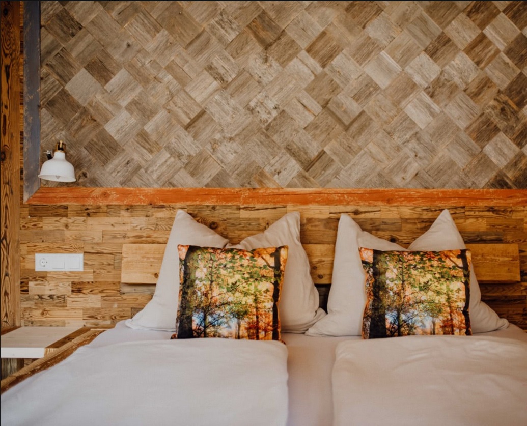 Gemütliches Doppelbett mit Rückwand aus Mareiner Altholz Gstaad und geflochtener Altholzverkleidung. Dekokissen mit Waldbild und warmem Licht schaffen eine naturnahe, entspannte Atmosphäre.
