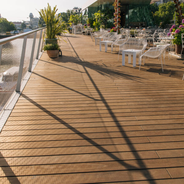 Close-up of the kebony wooden terrace of the Motto am Fluss restaurant with shadows, white furniture and floral decorations on the banks of the Danube.