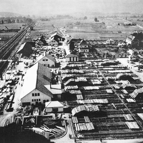 Black and white photo of a historic sawmill with large timber storage areas and adjacent railroad track - the Mattner sawmill as an early industrial complex in a rural setting.