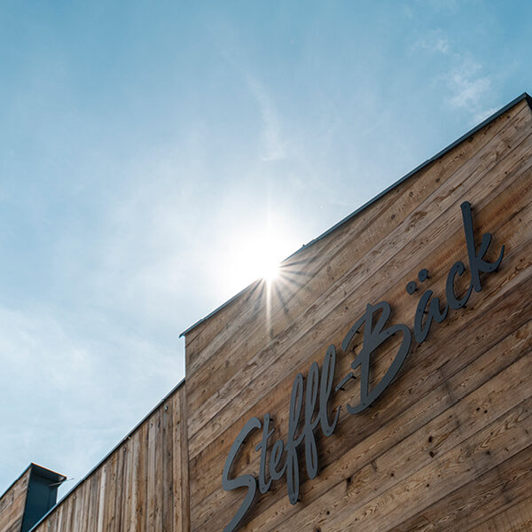 Moderne Holzfassade mit Schriftzug „Steffl-Bäck“, gefertigt aus Mareiner Holz. Die Sonne scheint hinter dem Gebäude hervor und erzeugt einen Lichtstern am Himmel.