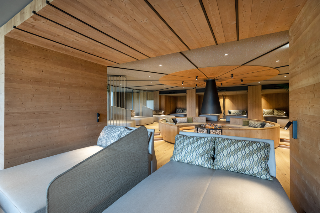Moderner Ruheraum mit stilvoller Holzverkleidung aus Mareiner Holz, gemütlichen Liegen und einem zentralen Kamin. Die warme Atmosphäre lädt zum Entspannen ein.