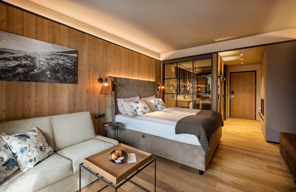 Gemütliches Hotelzimmer im Almwellness Hotel Pierer mit Wandverkleidung aus Mareiner Holz Zugspitze, modernem Interieur, Boxspringbett, Sofa und Glaswand zum Bad. Natürliche Materialien und warme Beleuchtung schaffen eine behagliche Atmosphäre.