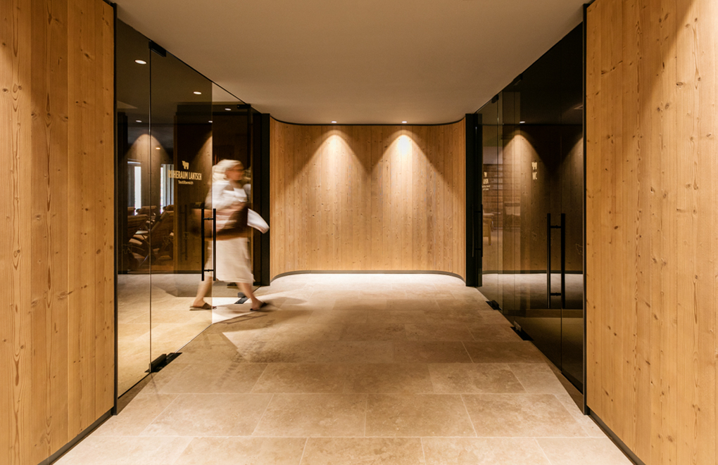 Eleganter Wellnessbereich im Almwellness Hotel Pierer mit Wänden aus Mareiner Holz Zugspitze, dezenten Deckenspots und Glasabtrennungen. Eine Person in Bademantel betritt den Raum, der eine warme, entspannende Atmosphäre ausstrahlt.