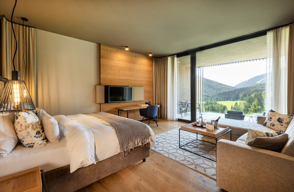 Modernes Hotelzimmer mit Panoramafenster, eleganter Einrichtung und Wandverkleidung aus Mareiner Holz Zugspitze. Gemütliches Bett, Sofa, Schreibtisch und Balkon mit Blick auf eine grüne Berglandschaft sorgen für Wohlfühlatmosphäre.