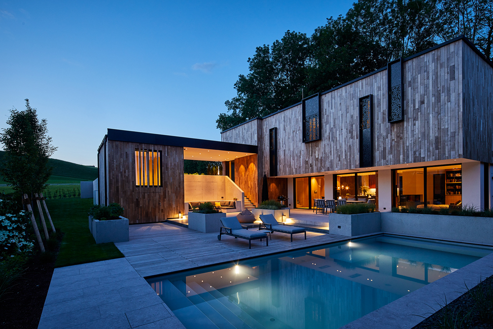 Abendaufnahme des modern gestalteten Privathauses Blaschke mit beleuchtetem Außenbereich, Pool und Holzfassade von Mareiner Holz. Große Fenster, stilvolle Ornamentfenster und elegante Außenmöblierung prägen das stimmungsvolle Ambiente.