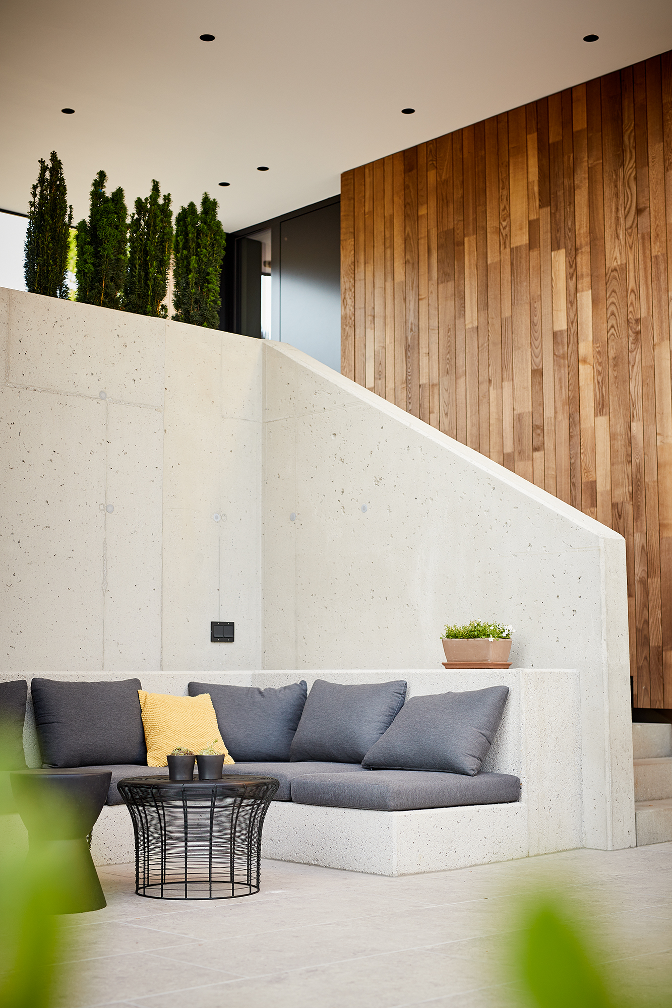 Nahaufnahme einer modernen Outdoor-Sitzgruppe im Privathaus Blaschke: Eckbank aus Sichtbeton mit grauen Polstern und gelbem Kissen, davor ein schwarzer Beistelltisch. Dahinter Sichtbetonwand mit integrierter Treppe und vertikaler Holzverkleidung.