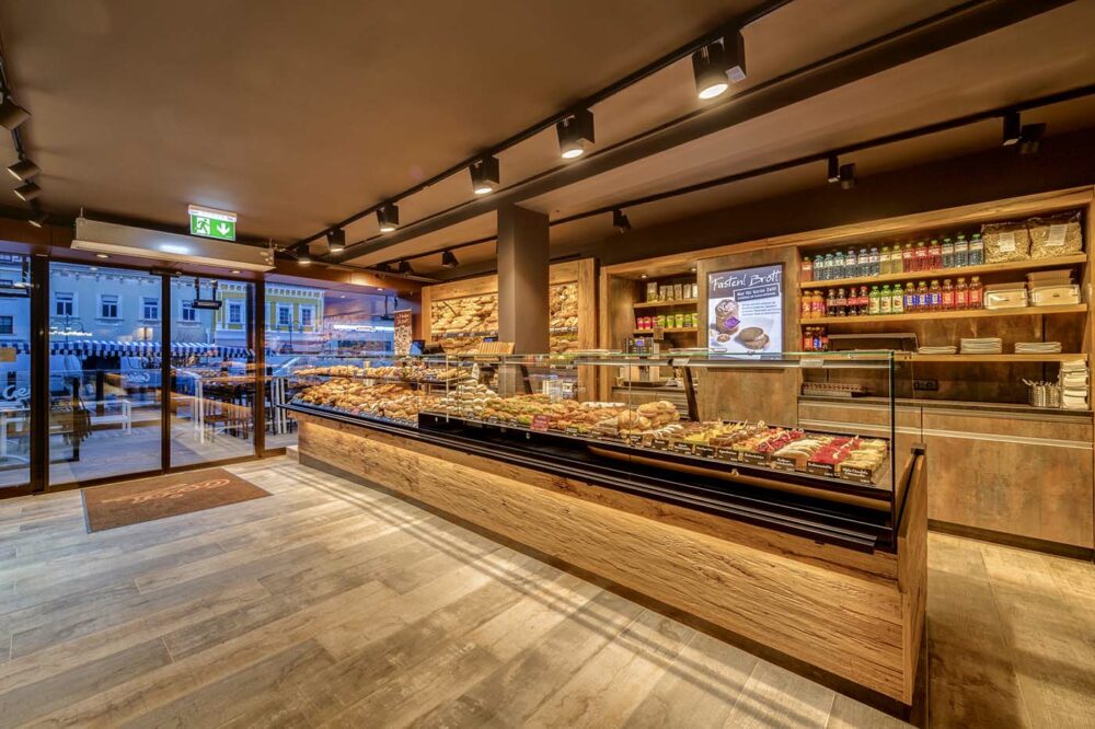 Heller Verkaufsraum der Bäckerei Geier mit großer Theke aus Mareiner Davos Holz, reich gefüllter Auslage und Blick nach draußen auf den Wochenmarkt.