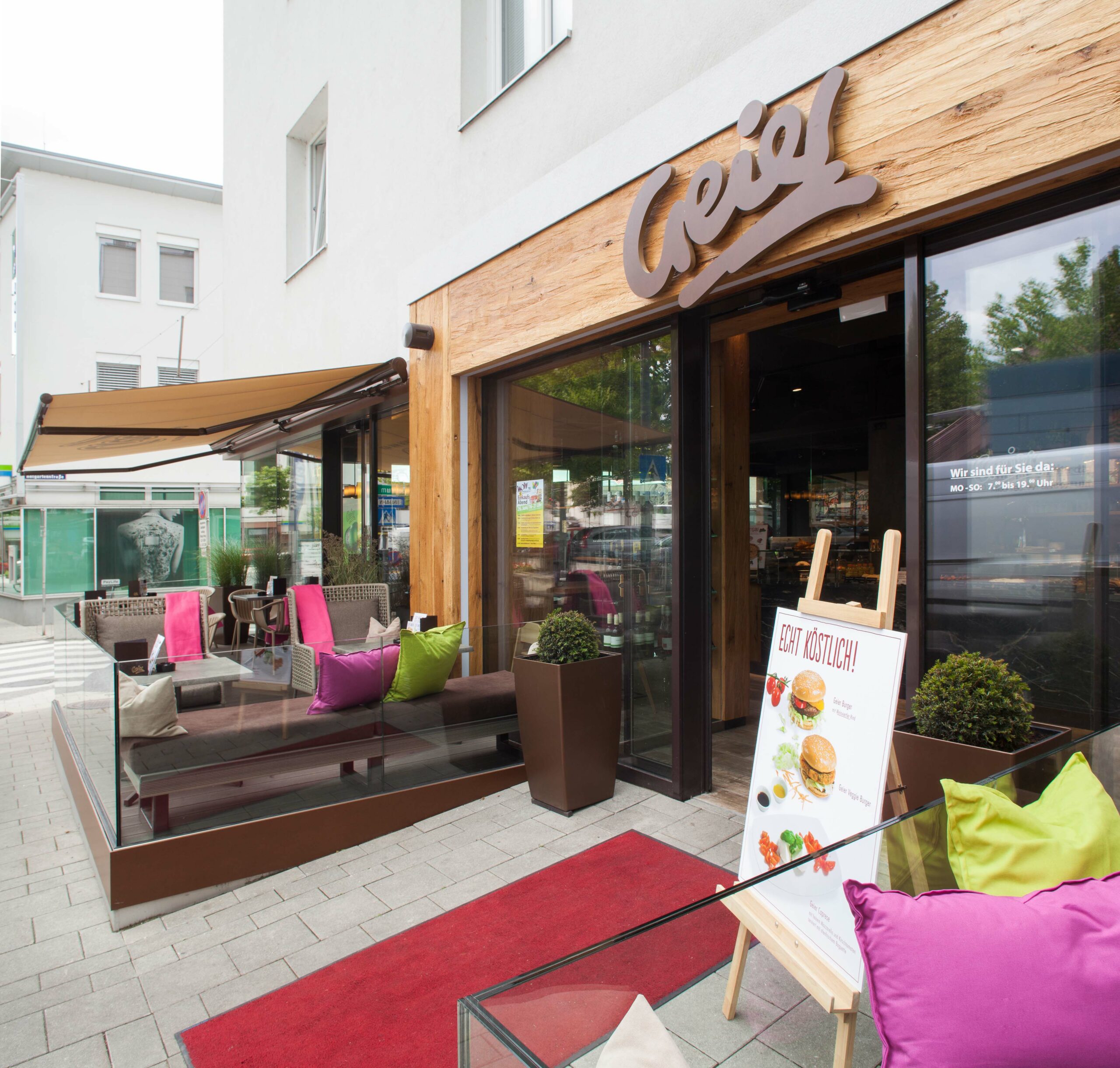 Außenansicht der Bäckerei Geier mit Holzfassade aus Mareiner Davos, gemütlicher Sitzlounge mit bunten Polstern und Werbetafel auf rotem Teppich – einladendes Ambiente in urbanem Umfeld.