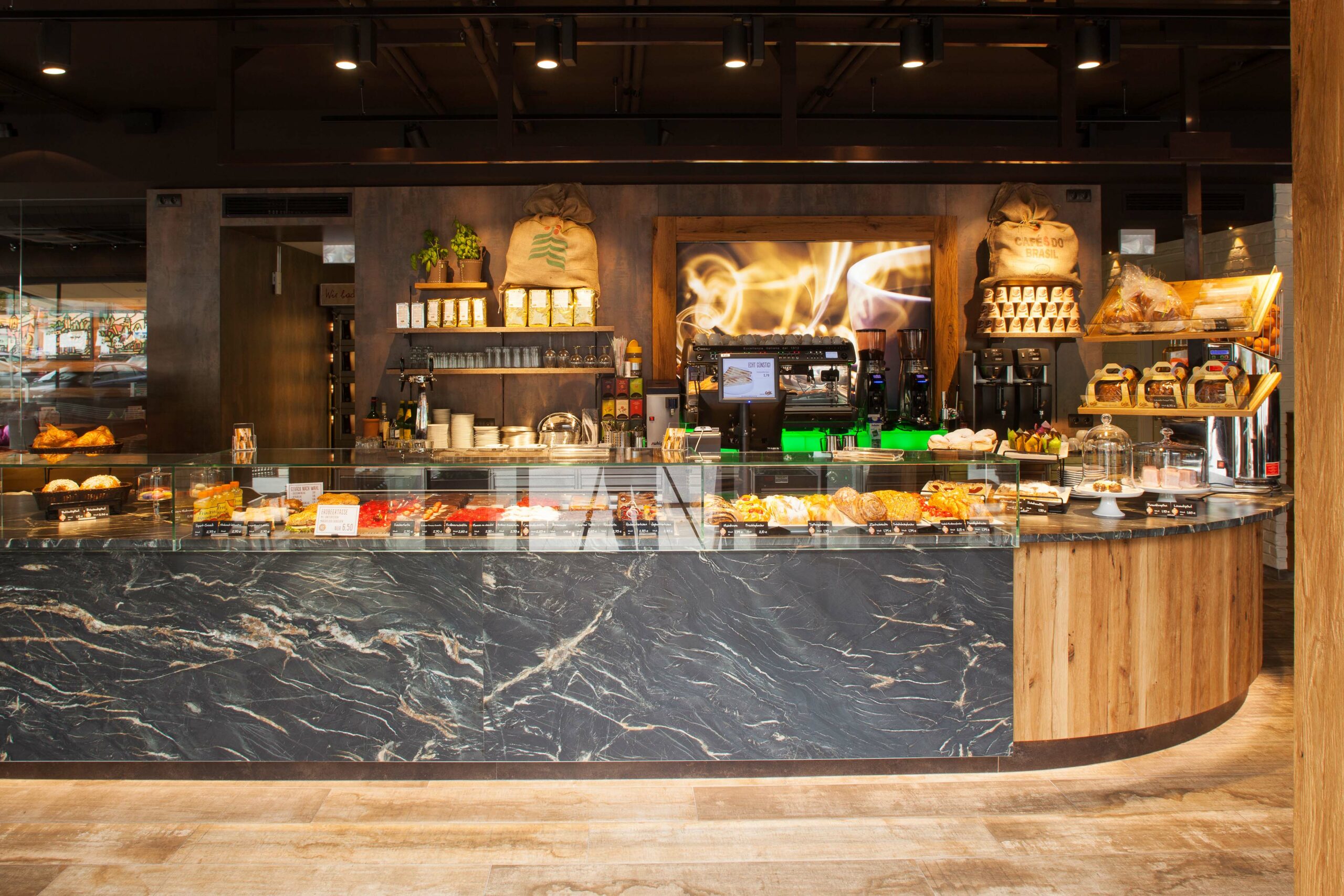 Frontalansicht der Bäckereitheke mit Steinverkleidung und Frontbereich aus Mareiner Davos Holz, dahinter Kaffeemaschinen, Gebäckauslage und stilvolle Kaffeehausatmosphäre.