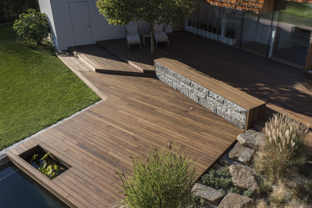 Großzügige Terrasse aus Kebony-Holz mit integrierten Ebenen, Natursteinmauer und eleganter Poolumrandung. Harmonisches Zusammenspiel von Holz, Stein und Grünflächen für eine stilvolle Outdoor-Oase.