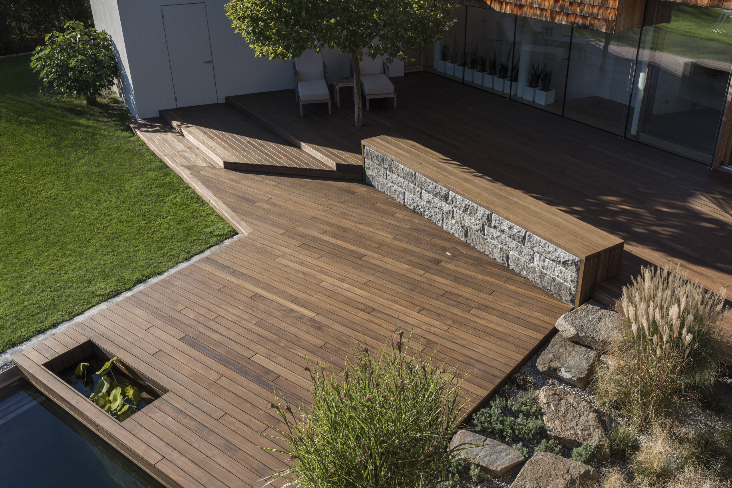 Großzügige Terrasse aus Kebony-Holz mit integrierten Ebenen, Natursteinmauer und eleganter Poolumrandung. Harmonisches Zusammenspiel von Holz, Stein und Grünflächen für eine stilvolle Outdoor-Oase.