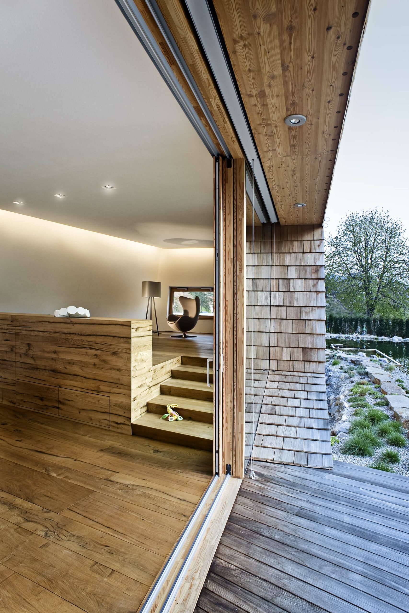 Blick aus einem modernen Wohnraum mit Holzboden und Verkleidungen aus Mareiner Simplon auf eine Terrasse aus Kebony-Holz. Sichtbar sind ein höher liegender Sitzbereich, ein Spielzeug auf den Stufen und der Garten mit Naturteich.