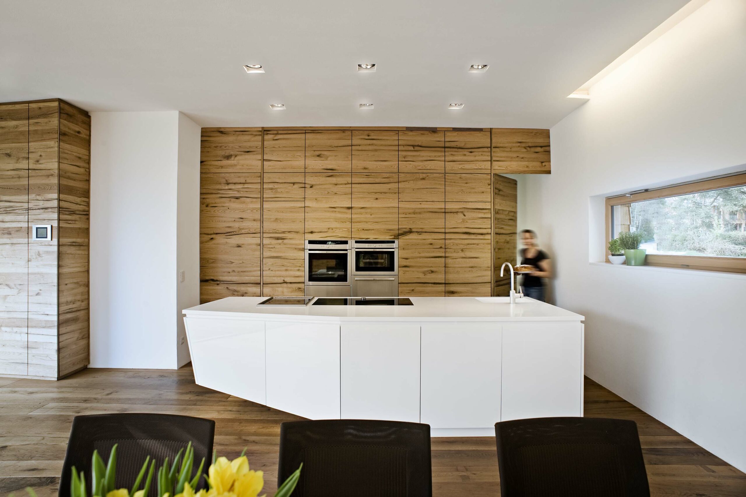 Helle, moderne Küche mit großem weißen Kochinselblock im Vordergrund. Die Rückwand ist mit hochwertigen Holzschränken aus Mareiner Davos verkleidet. Rechts ist ein Fenster mit Blick ins Freie, eine Person trägt ein Tablett.