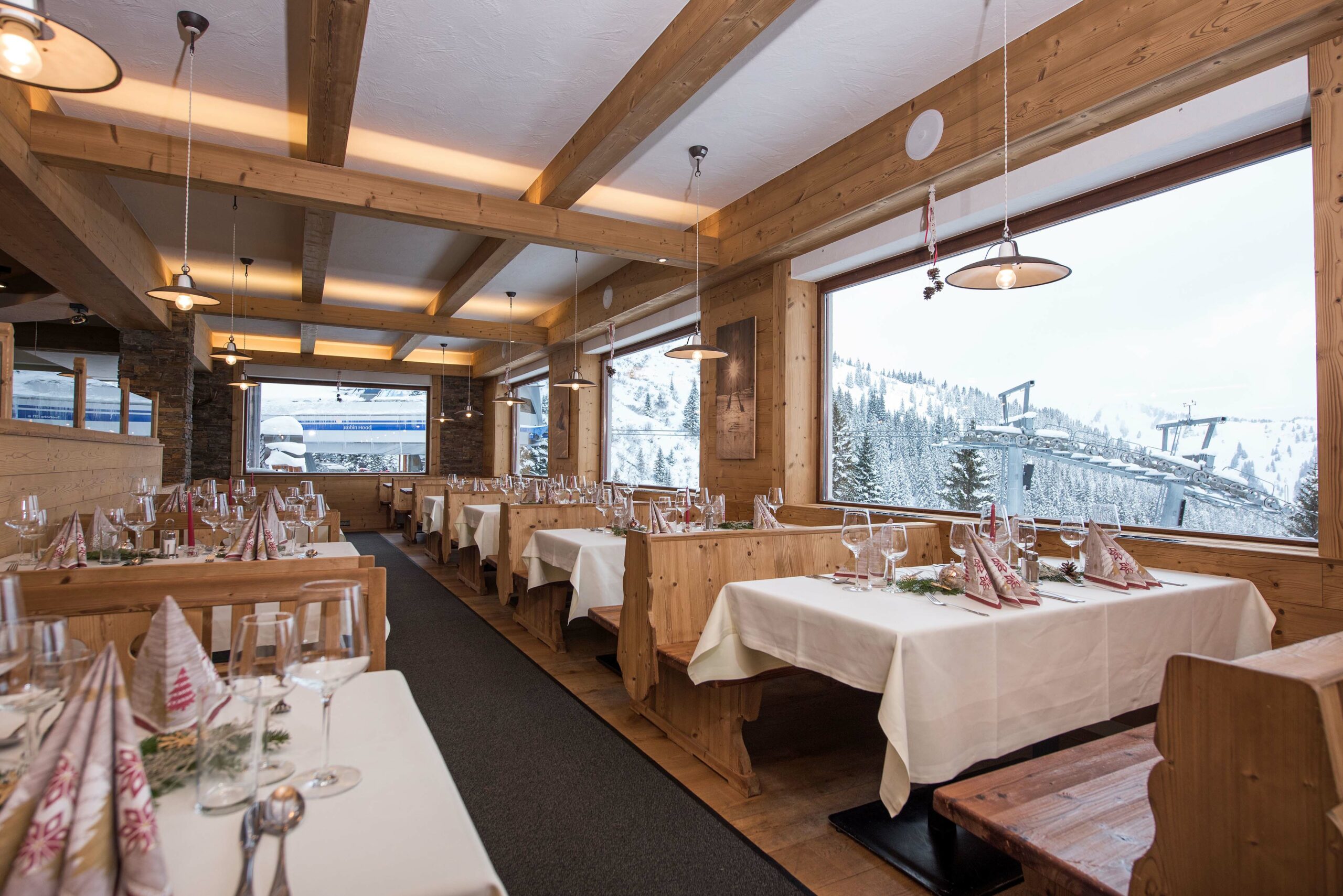 Gemütlicher Gastraum mit Holzverkleidung aus Gran Paradiso 190°, festlich gedeckten Tischen und Panoramafenstern mit Blick auf verschneite Pisten und Liftanlage.