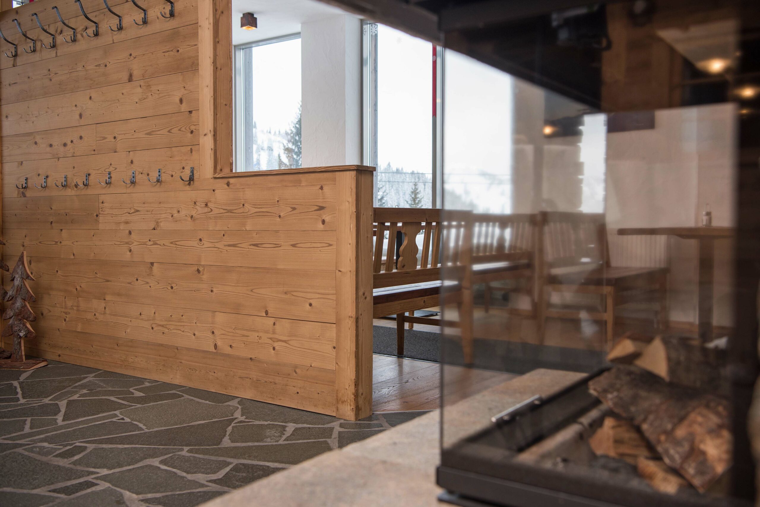Innenraum im Hotel Restaurant Berghof mit rustikaler Wandverkleidung aus Gran Paradiso 190° und traditionellem Mobiliar, ergänzt durch Natursteinboden und Blick auf die verschneite Winterlandschaft.