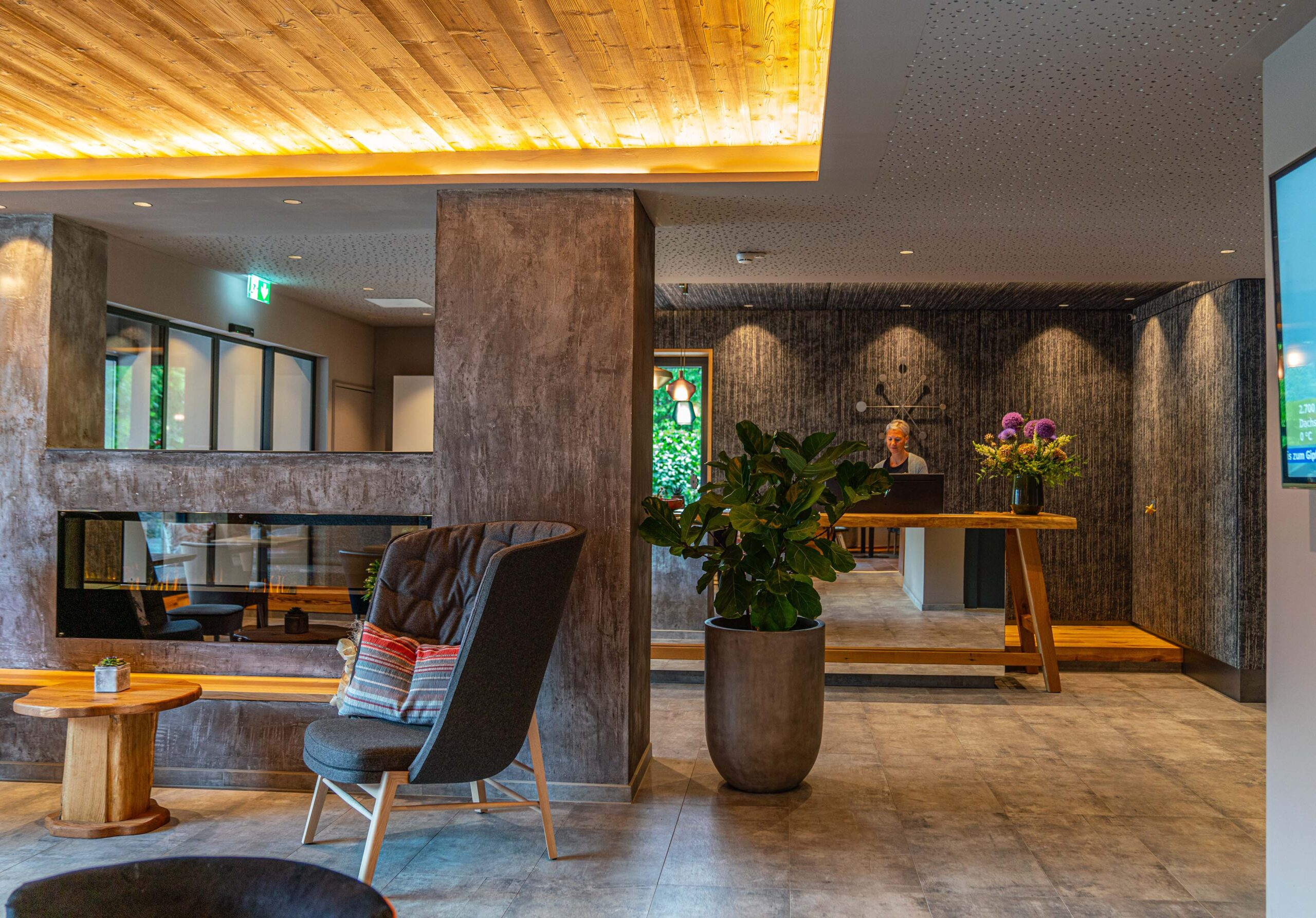 Moderne Hotellobby mit Empfangsbereich und gemütlicher Sitznische. Die Decke ist mit Mareiner Holz Marmolada Lava gestaltet, das für eine warme Atmosphäre sorgt. Akzente in Betonoptik und ein Kamin vervollständigen das elegante Design.