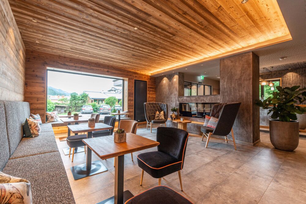 Heller, gemütlicher Loungebereich mit großen Panoramafenstern, Wand- und Deckenverkleidung aus Mareiner Holz Marmolada Lava. Moderne Sitzmöbel, warme Beleuchtung und Natursteinoptik schaffen eine einladende Atmosphäre.