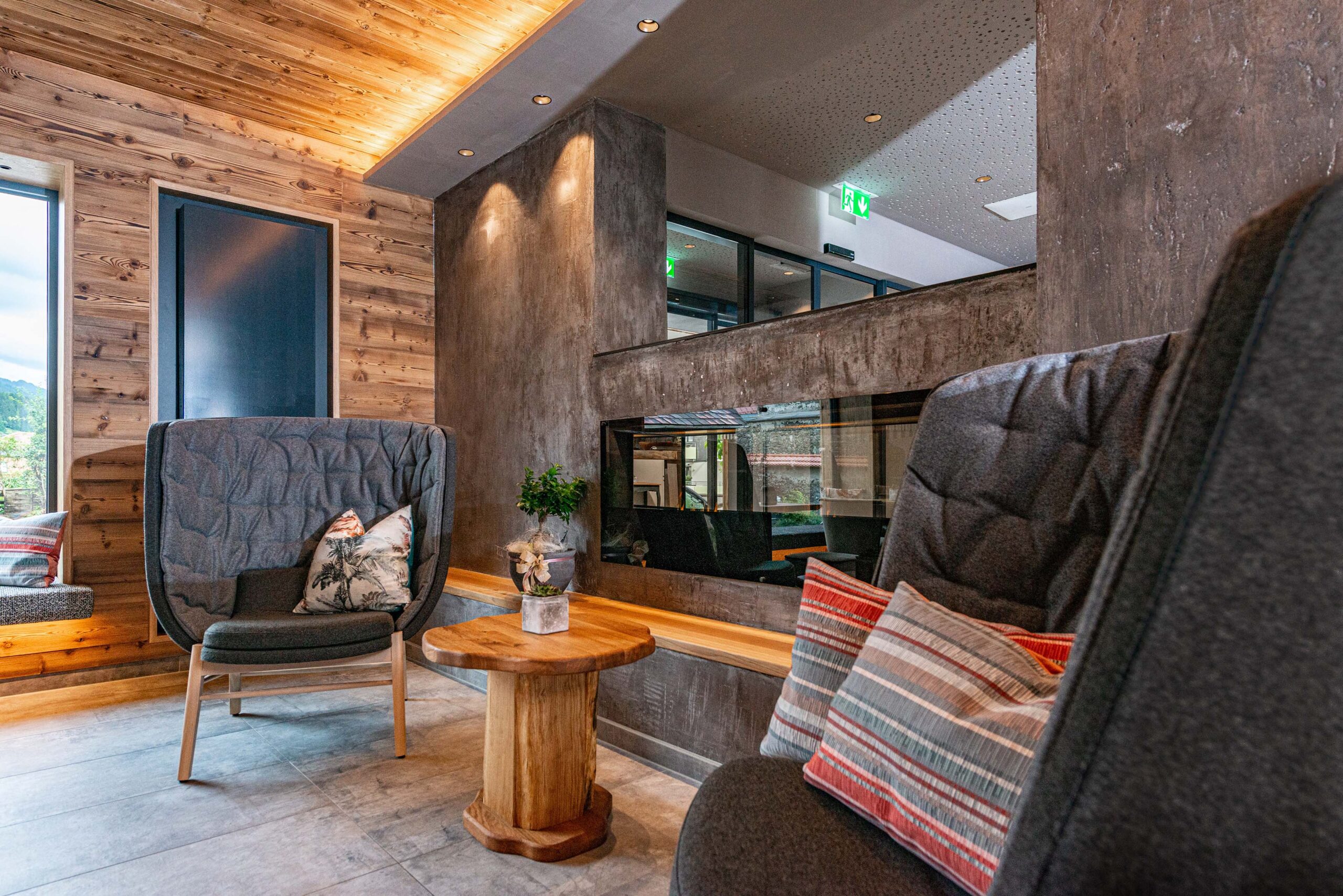 Ein moderner Loungebereich mit bequemen Sesseln, Natursteinoptik und Wänden aus Mareiner Holz (Marmolada Lava). Die warme Holzdecke mit Beleuchtung sorgt für Gemütlichkeit, während große Fenster natürliches Licht einlassen.