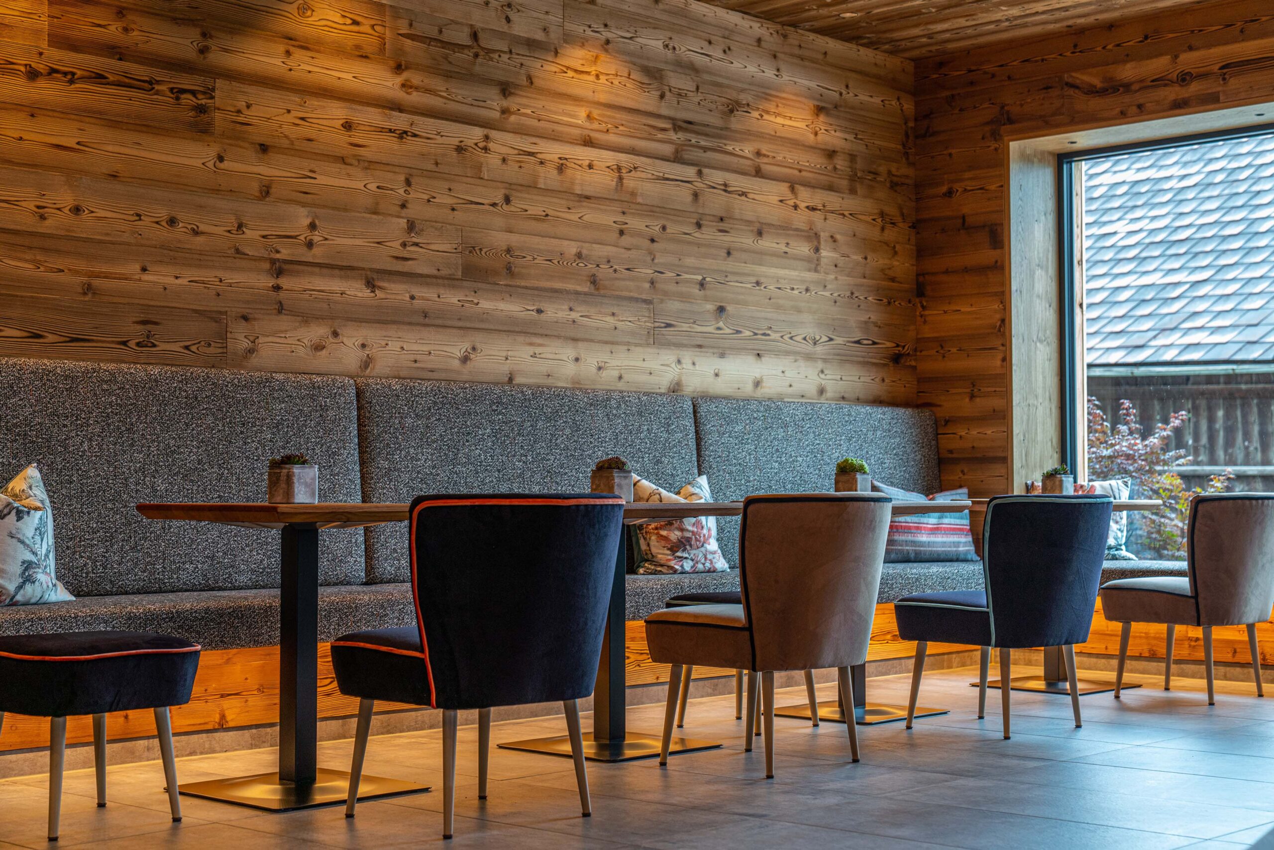 Ein stilvoll eingerichteter Gastraum mit Holzwänden aus Mareiner Holz (Marmolada Lava) und großen Fenstern. Gepolsterte Sitzbänke und elegante Stühle umrahmen schlichte Holztische. Warmes Licht schafft eine gemütliche Atmosphäre.