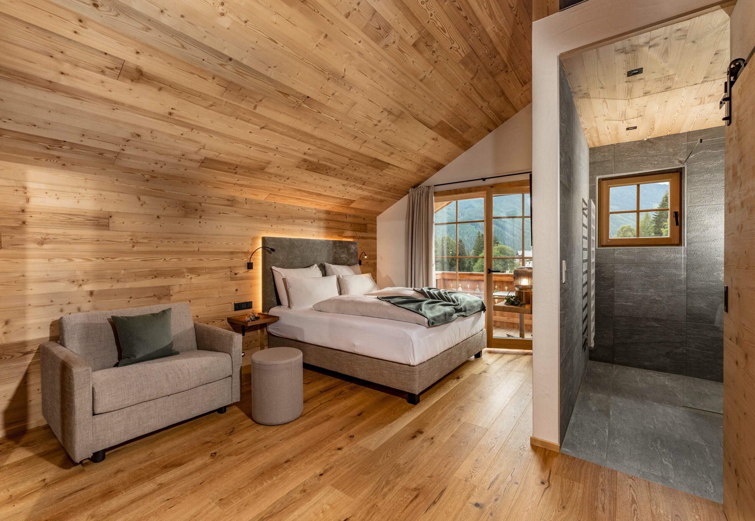Modern-alpines Schlafzimmer mit Decke und Wandverkleidung aus Marmolada-Holz. Im Raum befinden sich ein Doppelbett, ein Sofa und ein kleiner Hocker. Ein angrenzendes Bad mit Fenster und grauer Fliesenverkleidung ist offen zugänglich, im Hintergrund ein Bergblick durch die Glastür zum Balkon.