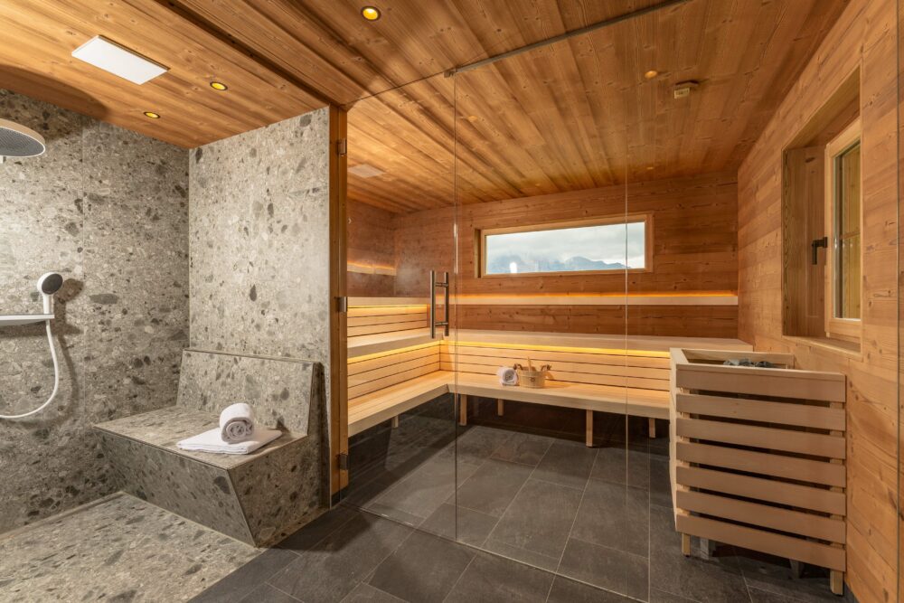 Moderne Sauna mit Wand- und Deckenverkleidung aus Gran Paradiso Holz. Im Vordergrund eine große Dusche mit Natursteinoberflächen. Die Sauna ist mit breiten Liegebänken, stimmungsvollem Licht und einem Fenster mit Bergblick ausgestattet.