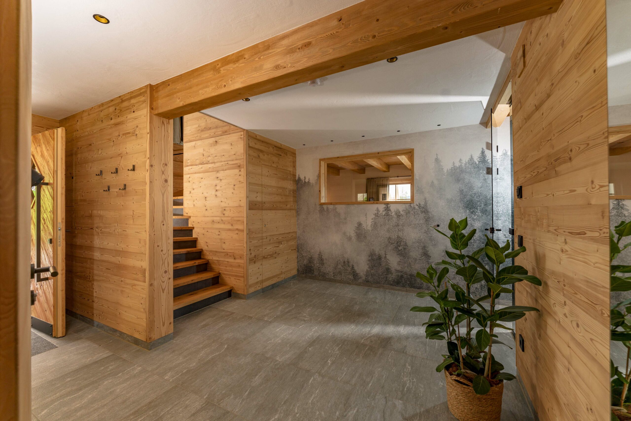 Moderner Eingangsbereich mit Wandverkleidungen und Türrahmen aus Marmolada-Holz. Eine Natursteinoptik-Fliese bedeckt den Boden, an der Wand ist ein stimmungsvolles Waldbild zu sehen. Eine Zimmerpflanze und sichtbare Holztreppe ergänzen das harmonische Ambiente.