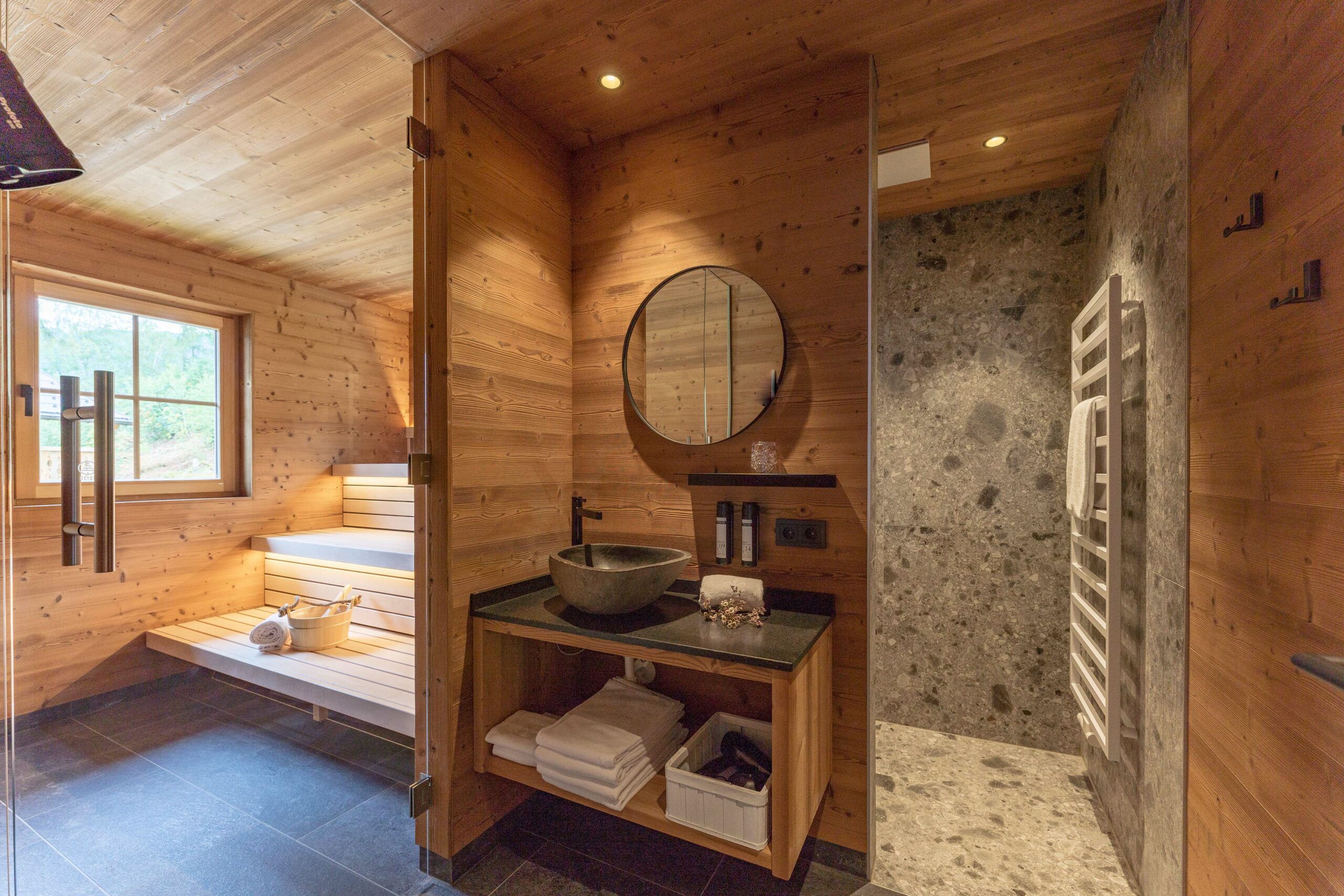 Hochwertiger Sauna- und Badbereich mit durchgängiger Wand- und Deckenverkleidung aus Gran Paradiso-Holz. Die Dusche ist mit Naturstein ausgekleidet, davor ein Waschtisch mit rundem Spiegel und schwarzer Armatur. Die Sauna bietet eine beleuchtete Sitzbank und ein Fenster mit Blick ins Grüne.