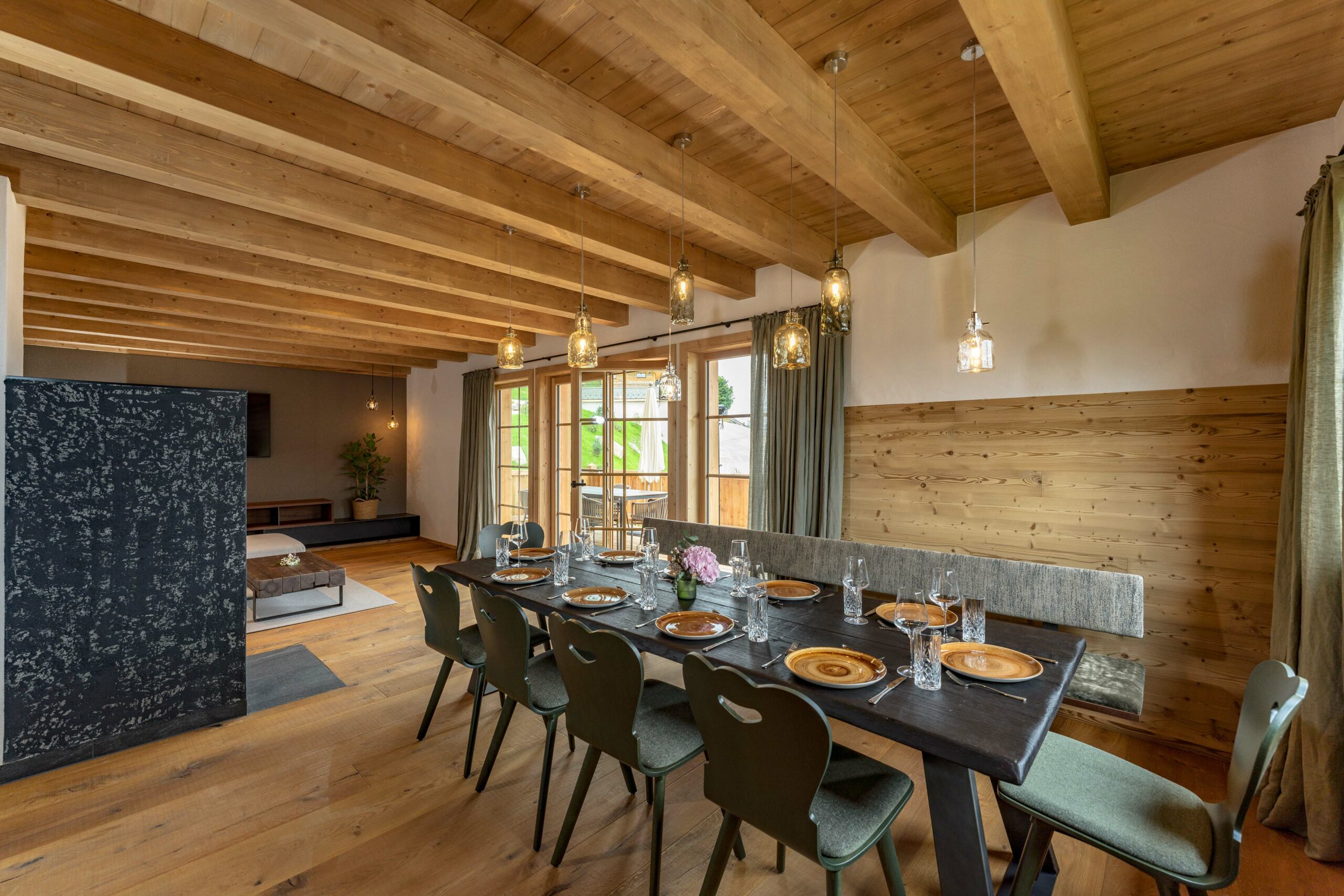 Gemütlicher Essbereich mit sichtbarer Balkendecke und halbhoher Wandverkleidung aus Gran Paradiso-Holz. Ein großer Esstisch ist für acht Personen gedeckt, darüber hängen stilvolle Glasleuchten. Durch große Fenster fällt Tageslicht in den offenen Wohnraum.