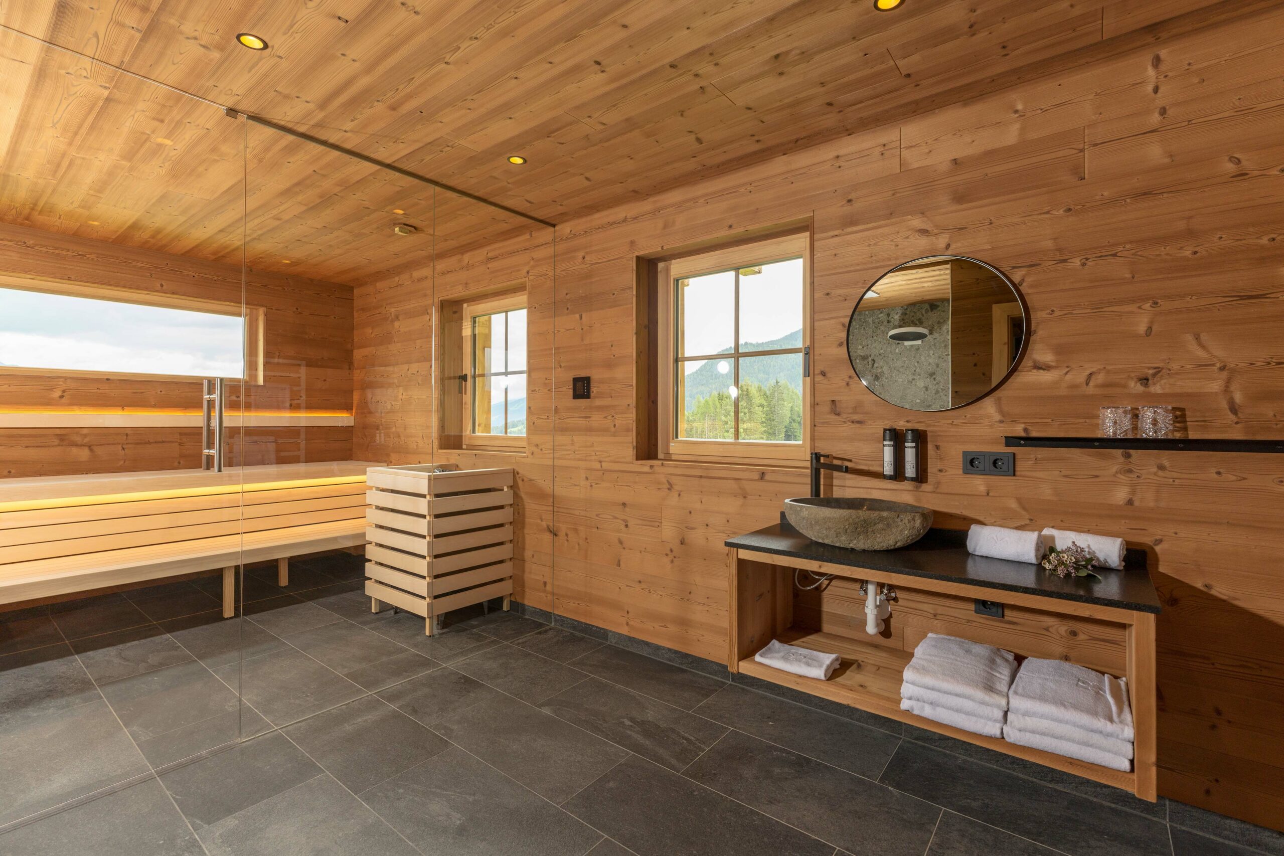 Großzügiger Wellnessbereich mit Wand- und Deckenverkleidung aus Gran Paradiso-Holz. Eine Glaswand trennt die Sauna mit beleuchteter Liegebank vom Waschbereich. Unter einem runden Spiegel steht ein Naturstein-Waschbecken auf einer Holzkonsole, daneben Fenster mit Blick ins Gebirge.