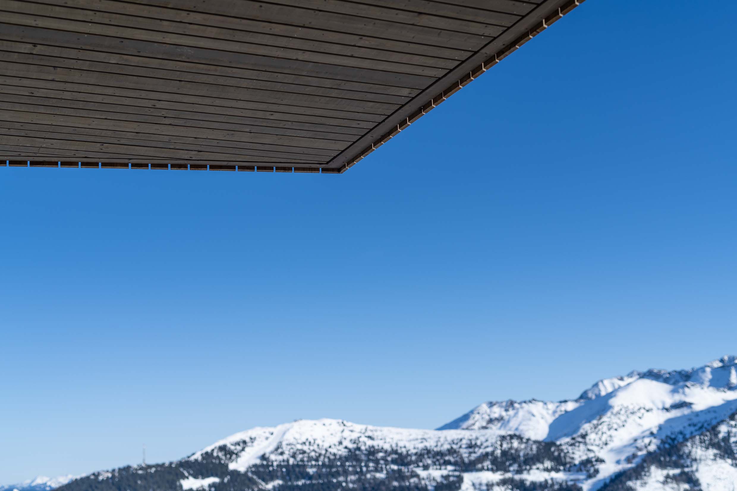 Minimalistische Nahaufnahme der Dachuntersicht der Planai-Bergstation, verkleidet mit Holzlamellen aus Mareiner Piz Nair. Die dunkle Holzstruktur kontrastiert mit dem tiefblauen Himmel, im Hintergrund schneebedeckte Gipfel und Pisten.