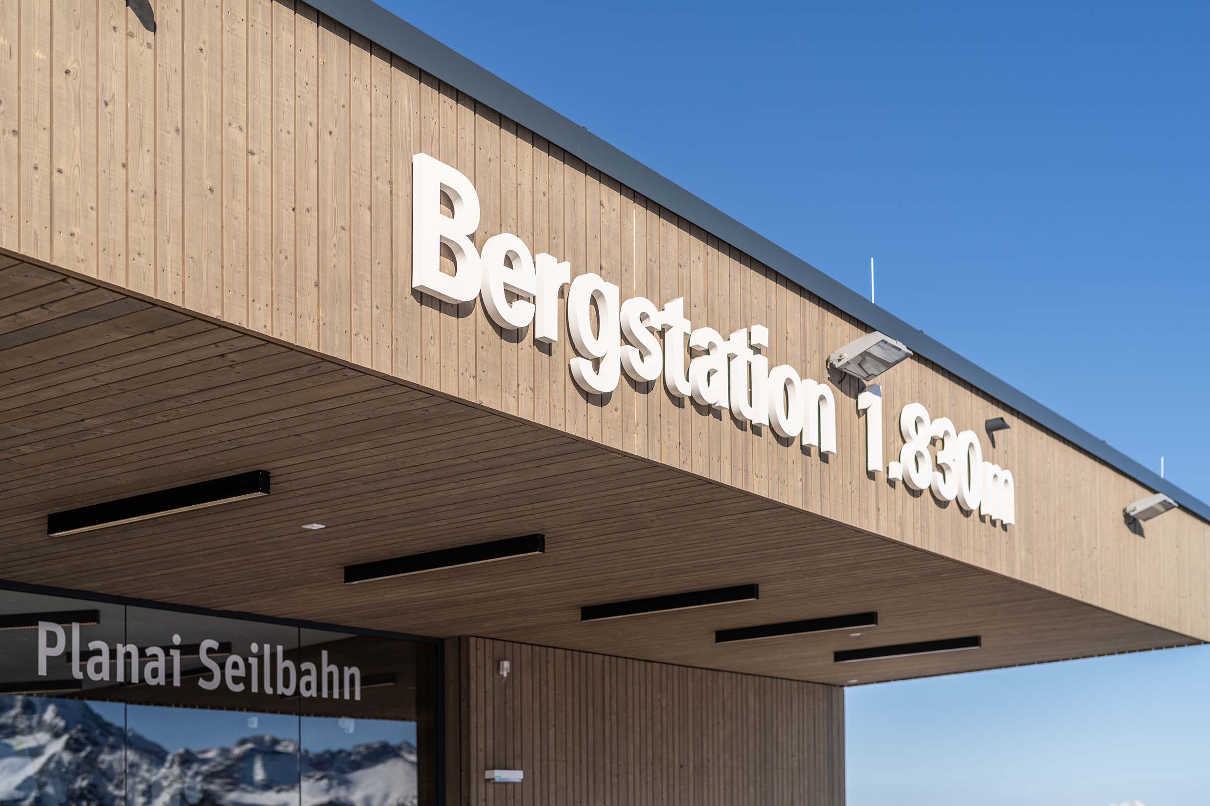 Nahaufnahme des Eingangsbereichs der Planai-Bergstation mit großem Schriftzug „Bergstation 1.830 m“. Die Fassade ist mit vertikalem Mareiner Piz Nair Holz verkleidet. Im unteren Bereich spiegelt sich die Bergkulisse in den Fenstern der Seilbahnstation.