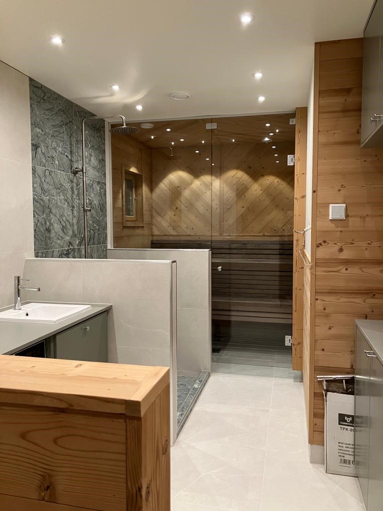 Modernes Bad mit begehbarer Dusche, Waschbecken und angrenzender Glas-Sauna in den Lappland Chalets, gestaltet mit Naturstein, Holzverkleidung und stimmungsvoller Beleuchtung.