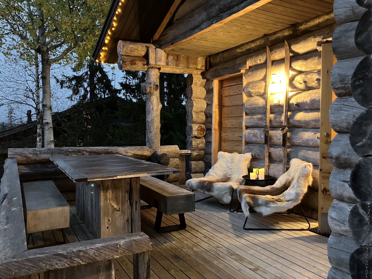 Gemütliche Holzterrasse der Lappland Chalets bei Dämmerung mit rustikalem Tisch, Bänken, zwei Sesseln mit Fellauflagen, Kerzenlicht und Blockhausfassade aus runden Baumstämmen.