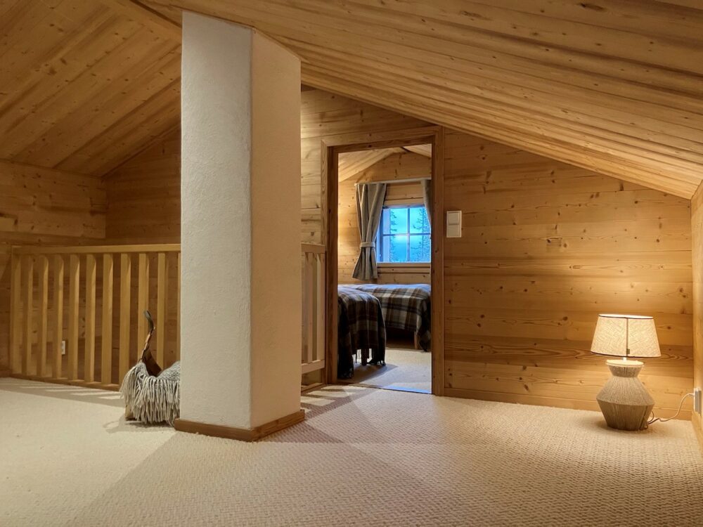 Gemütlicher Dachgeschossbereich der Lappland Chalets mit Holzwänden und -decke, Teppichboden, Stehlampe und Blick in ein Schlafzimmer mit karierten Textilien und Vorhängen.