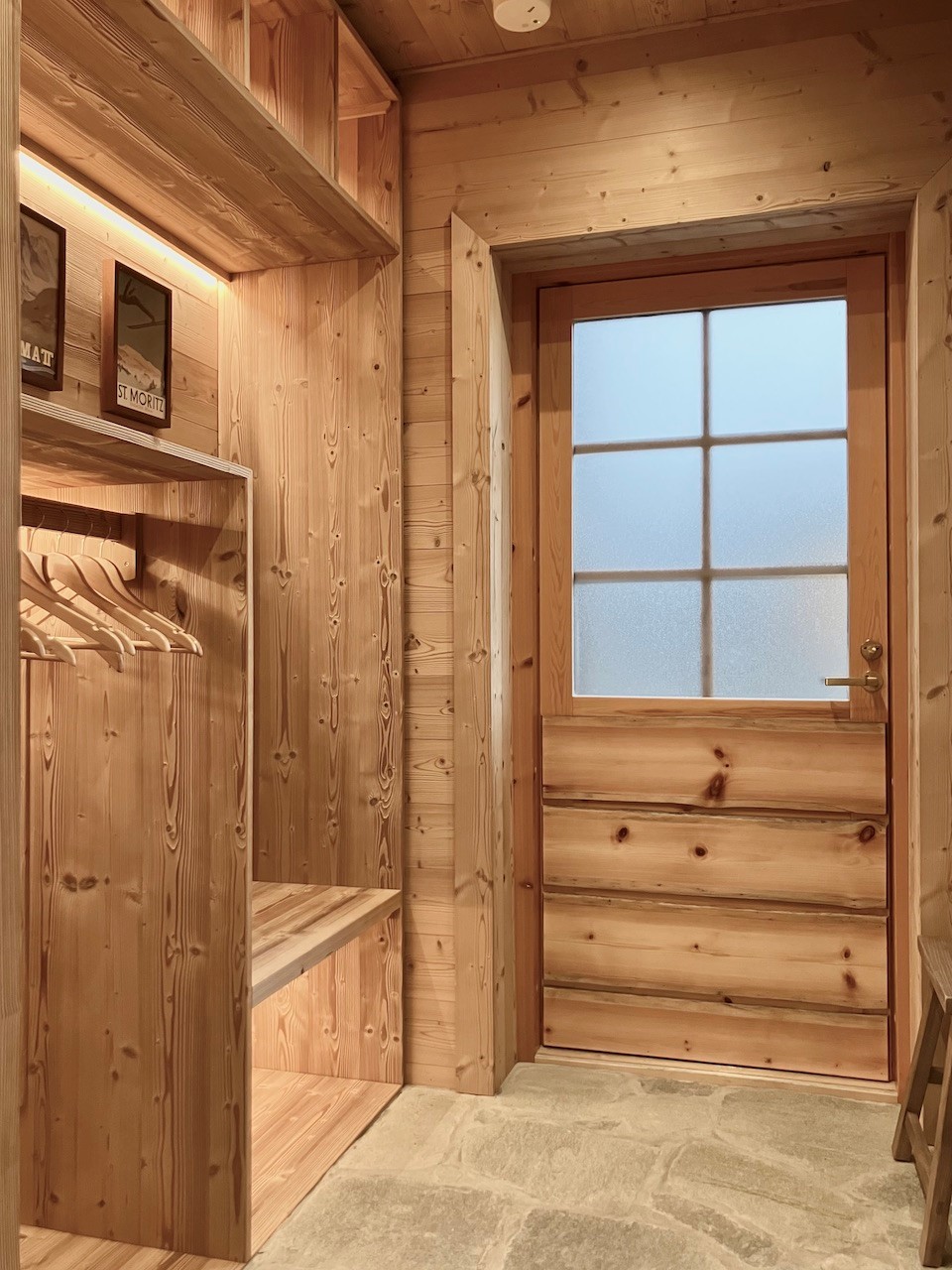 Heller Eingangsbereich der Lappland Chalets mit Holzverkleidung, Garderobe mit Sitzbank und Haken, dekorativen Skiplakaten und einer Holztür mit Milchglasfenster, kombiniert mit Natursteinboden.