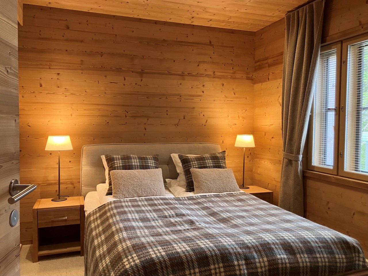 Gemütliches Schlafzimmer in den Lappland Chalets mit Holzverkleidung, Doppelbett, karierten Textilien und warmem Licht durch zwei Stehlampen sowie Tageslichtfenster mit Vorhängen.