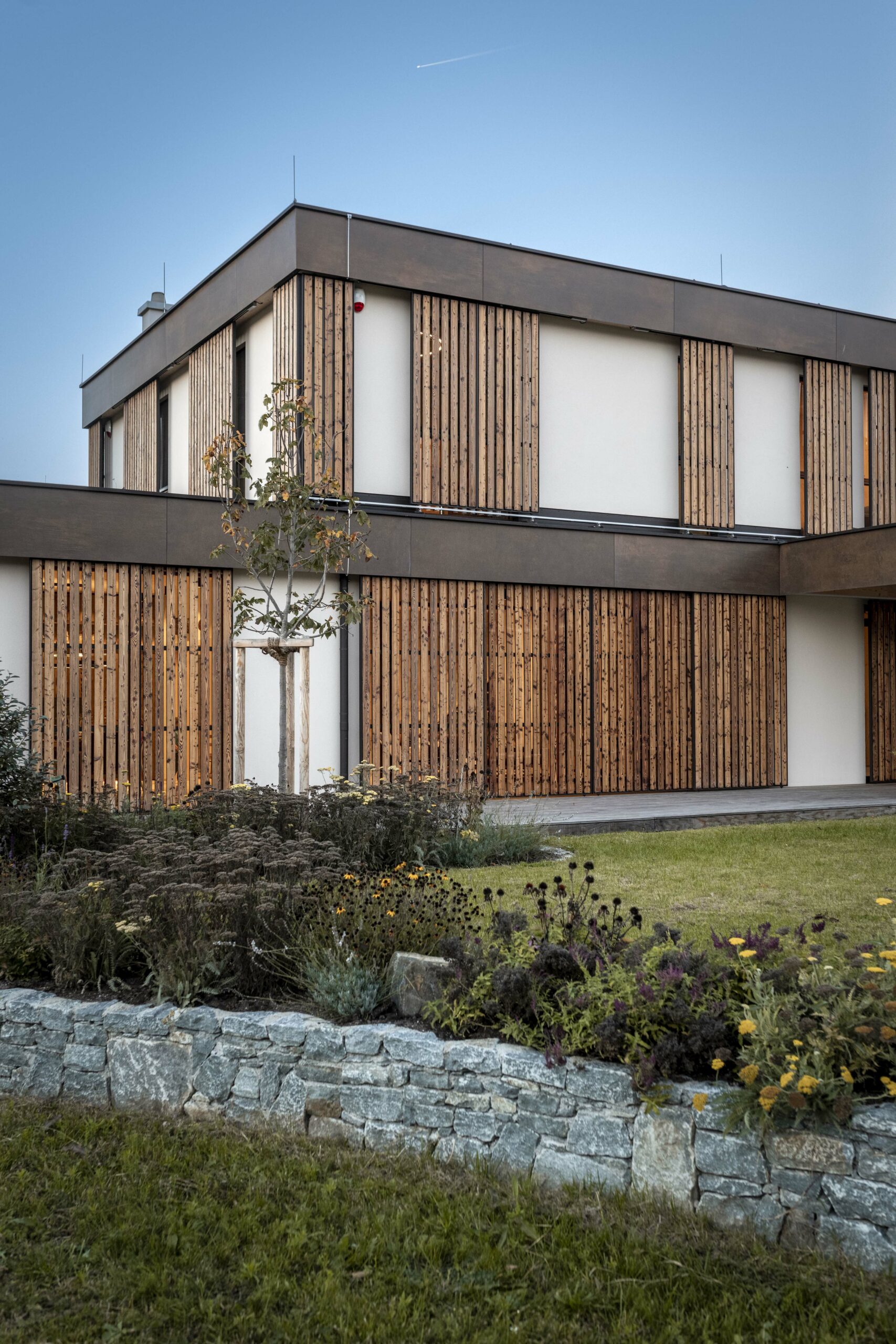 Fassade eines modernen Einfamilienhauses mit vertikalen Holzlamellen aus Piz Bernina Lava, kombiniert mit weißen Wandflächen und dunklen Rahmen. Im Vordergrund blühender Garten mit niedriger Natursteinmauer.